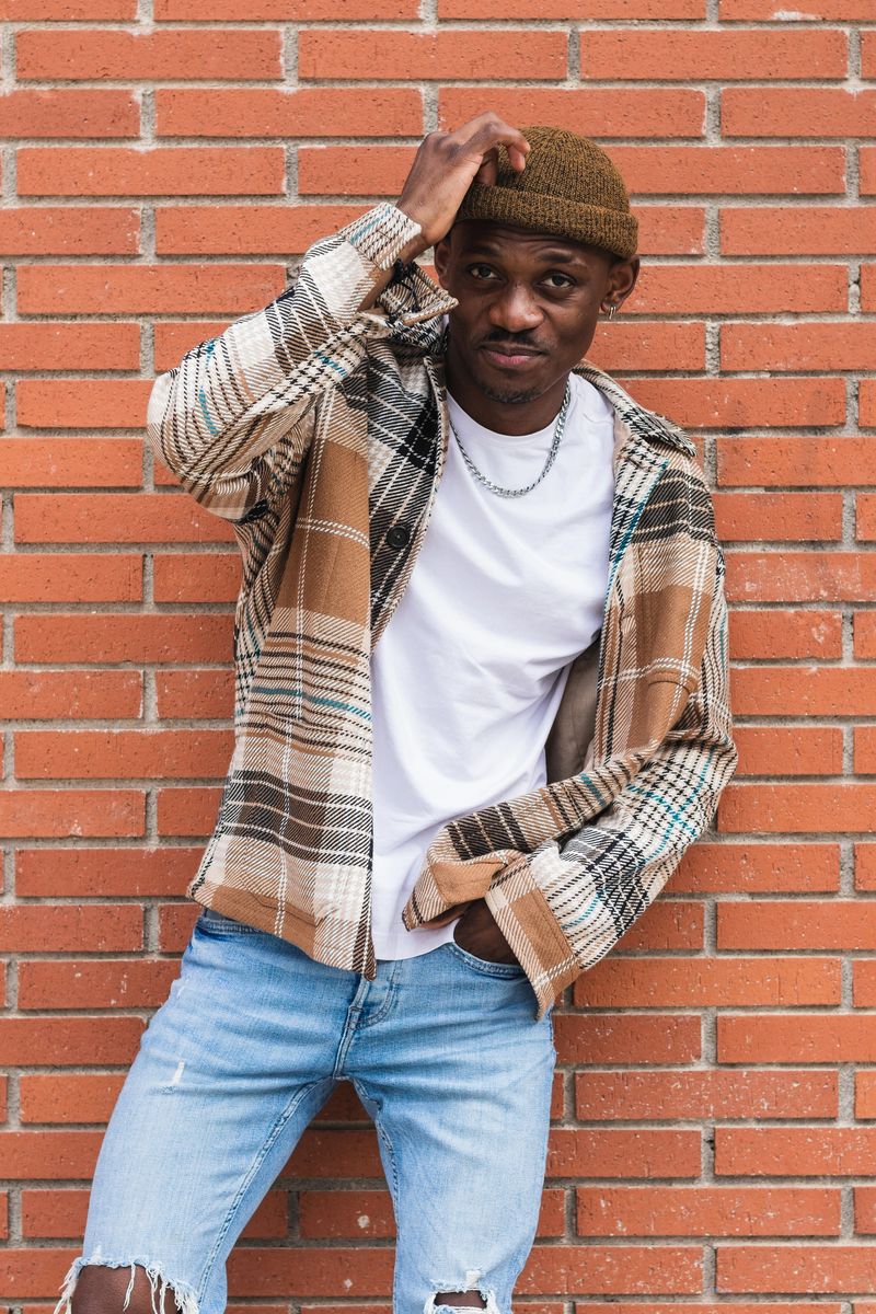Young African American guy in checkered shirt and trendy hat frowning and looking at camera while standing against brick wall on city street