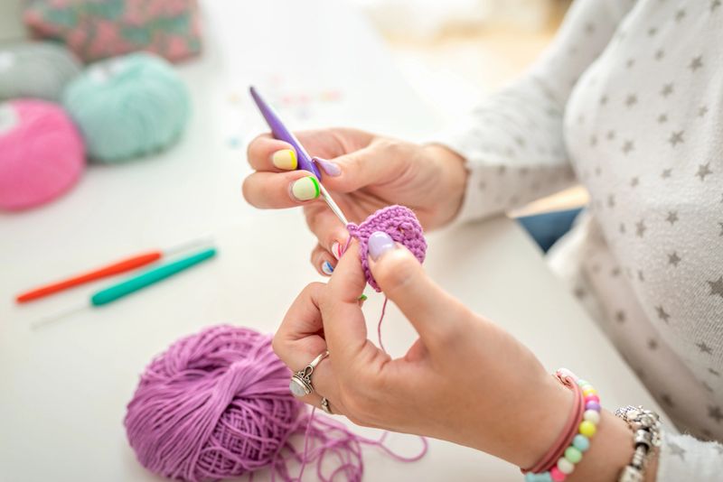 A close-up view of a woman’s hands as she skillfully crochets with purple yarn. The background includes colorful crochet hooks, balls of wool, and a pincushion, showcasing an organized and vibrant crafting environment. The bright workspace highlights her focus and creativity.