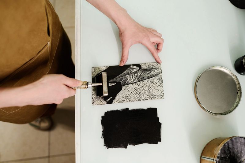 Top view of hands of young unrecognizable female designer or worker of printing house applying ink with paintroller on artwork