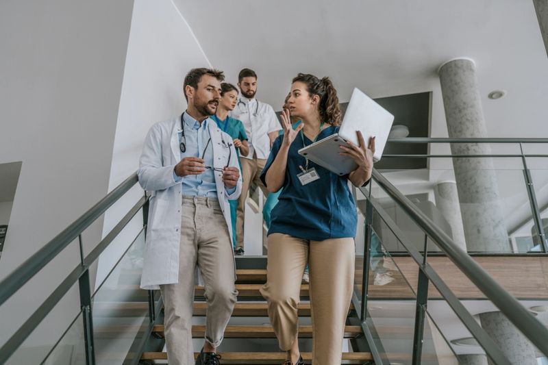 A group of doctors, dressed in white coats, walk up a set of stairs in a modern hospital building. One of the doctors, a woman, carries a laptop. The doctors appear to be engaged in conversation, perhaps discussing a patient or recent developments.