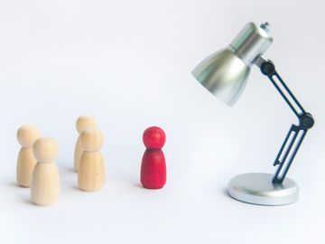A red wooden figure stands apart from four natural wood figures under a desk lamp.