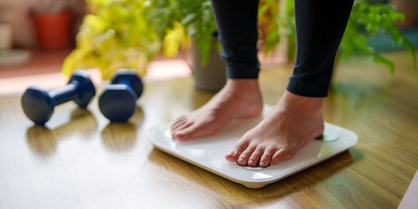 Person standing barefoot on a scale with dumbbells nearby.