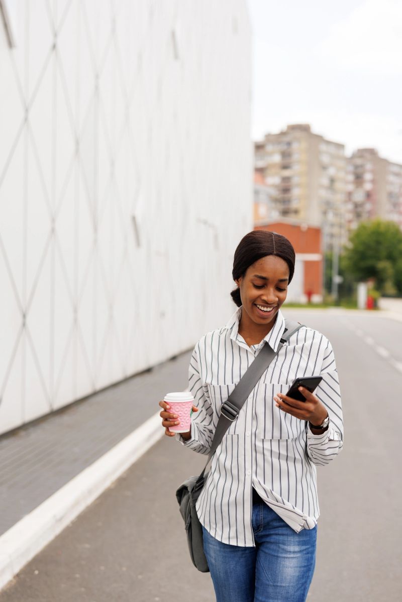 Young African businesswoman with laptop bag and take away coffee scrolling through social media on mobile phone in front of modern office building
