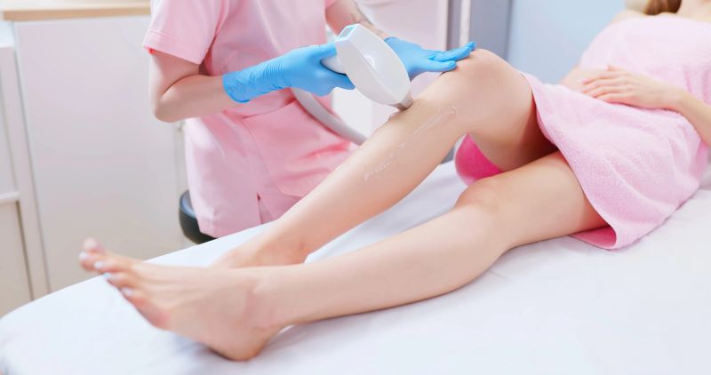 side view asian woman lying on bed is receiving leg hair removal procedure with laser machine while female doctor sit holding cosmetology device in clinic