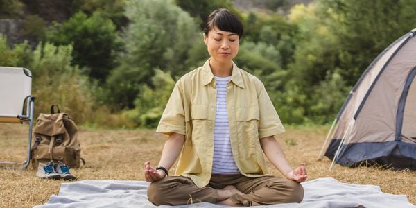 A woman meditates outdoors next to a tent and camping gear.