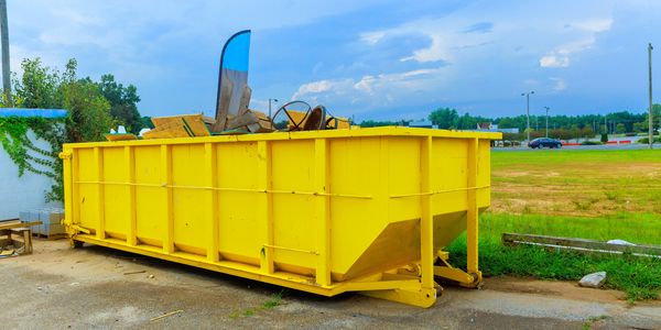 A large yellow dumpster filled with wooden debris sits outdoors under a cloudy sky.