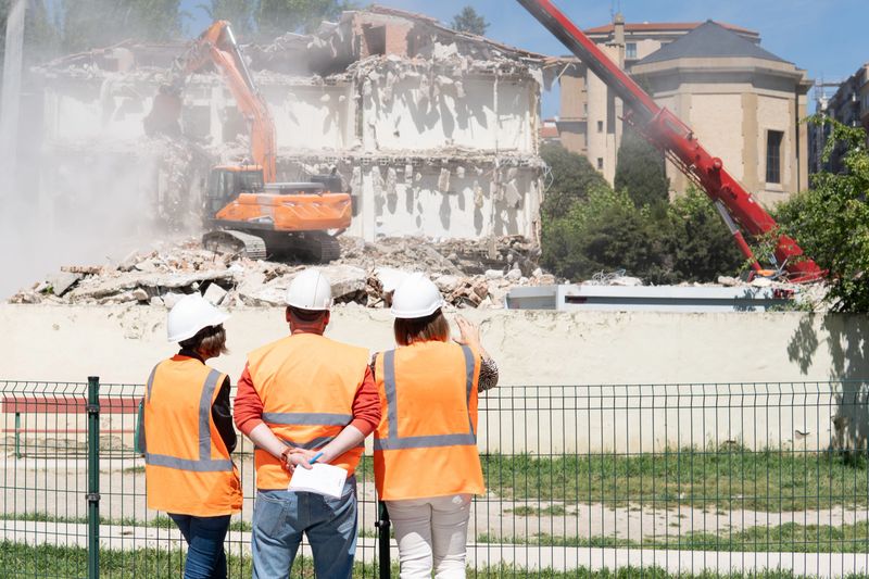 three professional engineers observe and point out the demolition of a house with an orange excavator, dressed in work clothes, helmet and vest