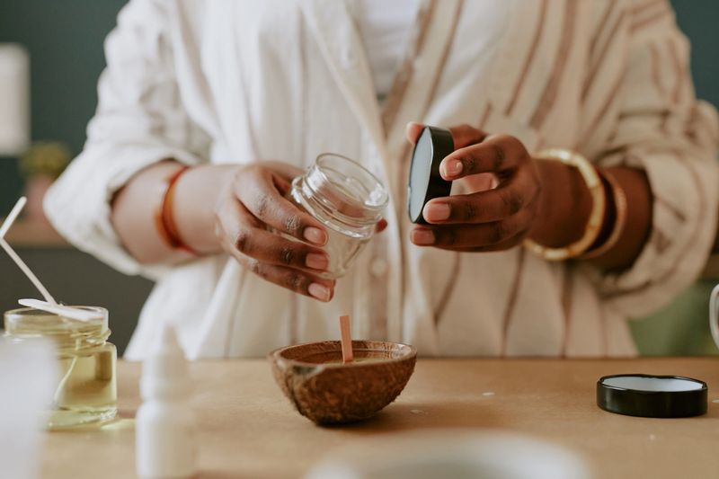 Woman preparing natural skincare product by pouring essential oil into a glass jar and mixing with ingredients on a wooden table, showcasing homemade beauty care process