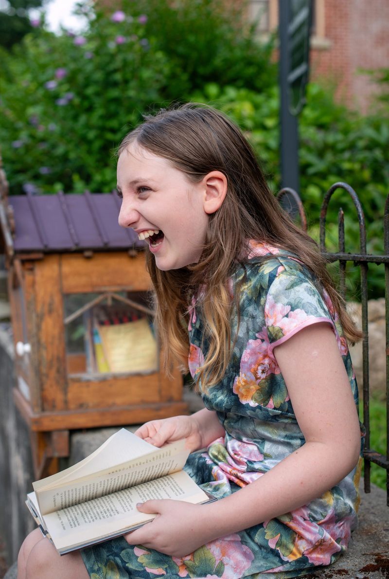 Cheerful girl in floral dress sits on stone ledge, engrossed in a book. Long brown hair frames her face as she reads in a lush garden setting. Wooden structure resembling a little free library stands in background. Scene captures simple pleasure of outdoor reading on sunny day, emphasizing joy of literature for young minds.
