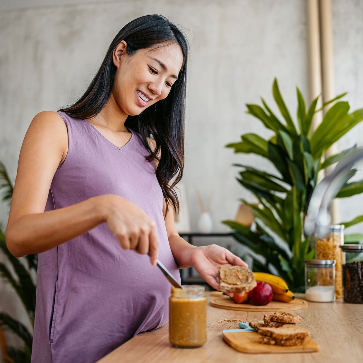 Pregnant woman spreading peanut butter on bread in a bright kitchen.