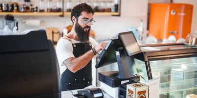 Bearded barista using a touchscreen register in a cozy café.