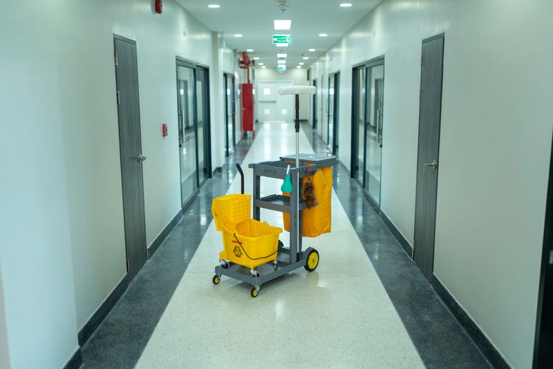 A yellow cleaning cart is in the middle of a hallway. The cart is empty and has a yellow bucket on it