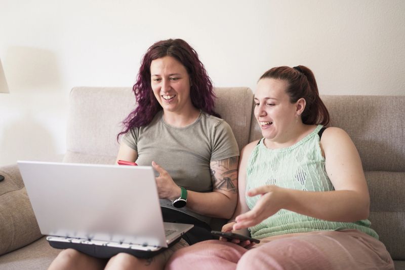 Two women sitting on a couch, one of them is using a laptop. They are laughing and smiling at something on the laptop