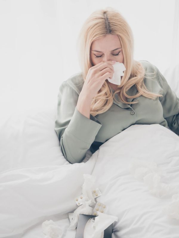 Woman in bed blowing her nose with tissues and medicine nearby.