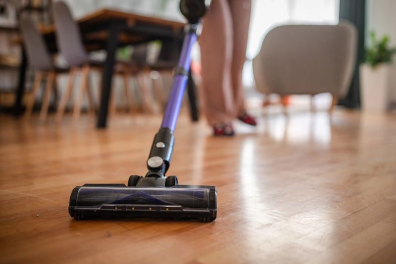 A close-up shot of a vacuum cleaner head in action on a wooden floor, reflecting cleanliness and maintenance. Furniture and home setting are slightly visible in the background.