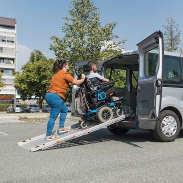 Woman pushing man in wheelchair up ramp into van.