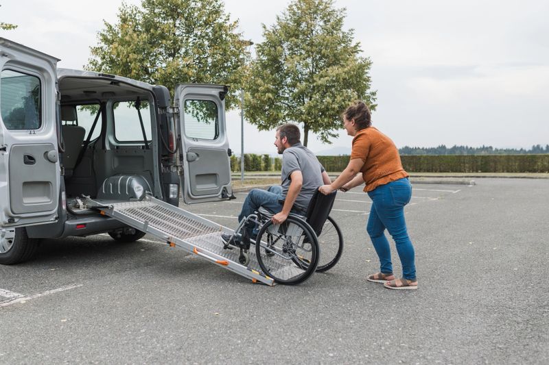 Female assistant helping a male person in wheelchair with transport using accessible vehicle van ramp. Disability and mobility concepts.