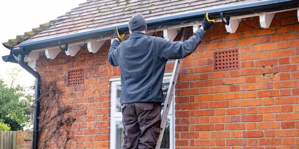 Man on ladder cleaning gutters of a brick house.