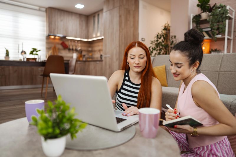 In a bright kitchen nook, two friends share ideas while working together on their laptops. One suggests notes as the other types, creating a warm atmosphere of creativity.