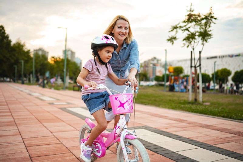 A girl is learning to ride her pink bike while her mother assists her in a lush park during a bright afternoon. They share smiles and encouragement as she gains confidence.
