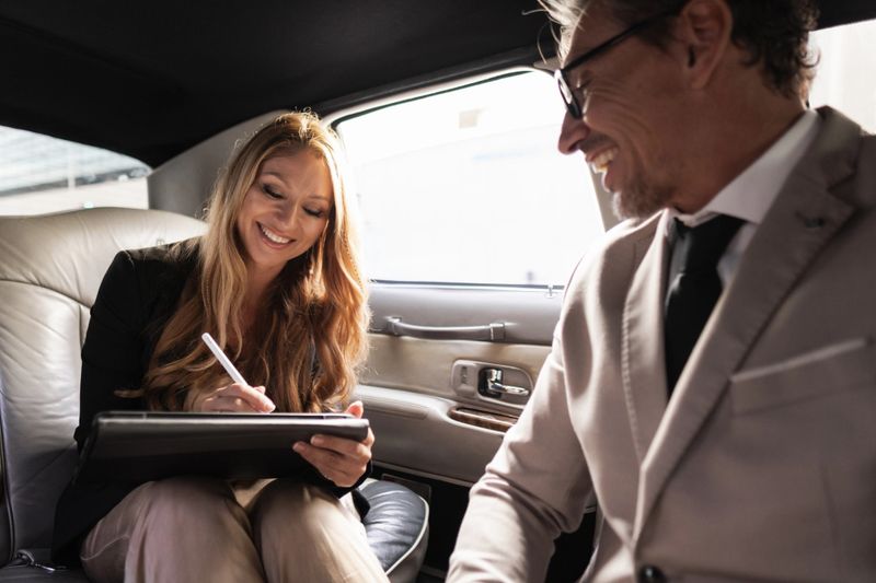 Businesswoman signing a contract on a digital tablet in the back of a limousine, with her business partner looking on