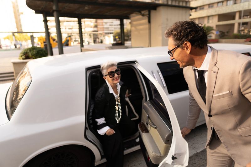 Elegant senior businesswoman is exiting a limousine with the help of her chauffeur in the city