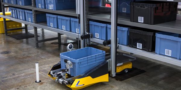 An autonomous robot carries a blue bin in a warehouse aisle.