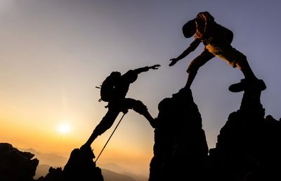 Two climbers helping each other reach the summit at sunset.