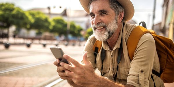 Smiling older man with a hat and backpack using a smartphone outdoors.