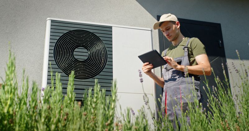 A young technician in overalls and a cap uses a tablet to inspect an outdoor heat pump unit installed on the exterior wall of a building. The scene is set on a sunny day, with the technician surrounded by lavender plants, adding a touch of nature to the industrial setting. The image emphasizes the integration of technology and nature in modern energy solutions.