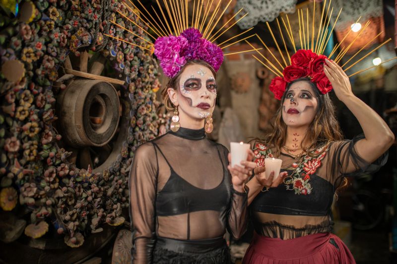 Mexican women dressed as Catrina with a headdress with flowers on their heads, alluding to the Day of the Dead, with a candle in their hands, looking up and to the side.