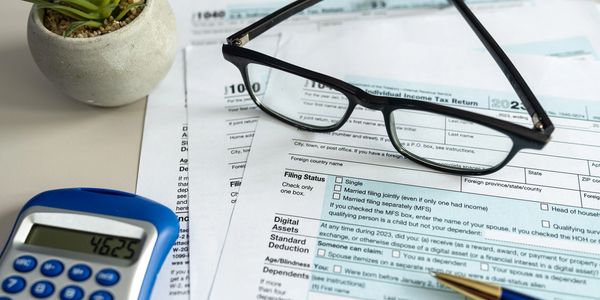 Tax forms with glasses, pen, and calculator on a desk near a small potted plant.