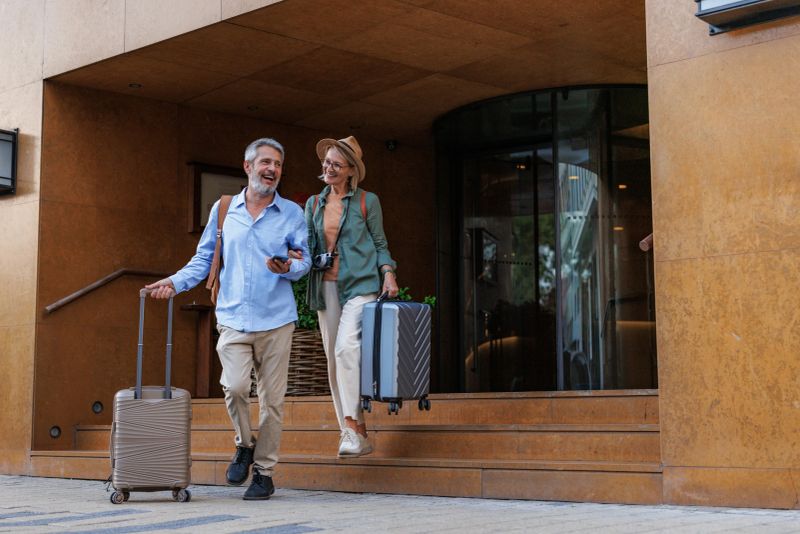 Mature couple joyfully exits a hotel, rolling their suitcases. They appear cheerful and ready for travel adventures. A sense of excitement and vacation spirit is captured in this moment.