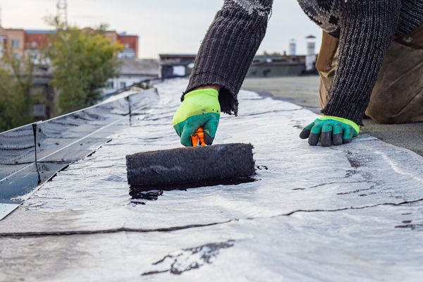 Worker applying black liquid sealant to a flat roof with a roller for waterproofing.