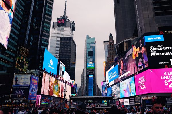Crowded Times Square with bright digital billboards and skyscrapers under an overcast sky.