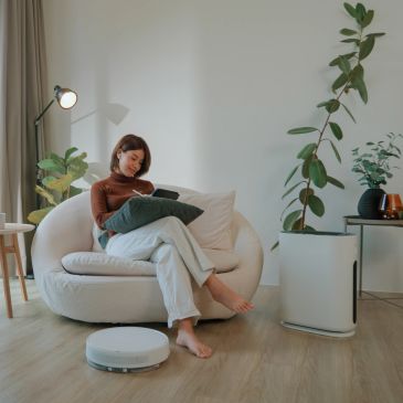Woman sitting on white sofa using phone with a robot vacuum in front.