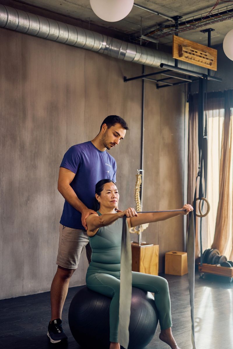 Physiotherapist guiding female patient through resistance band exercise on exercise ball. Full-body shot in rehabilitation clinic. Healthcare and physical therapy concept