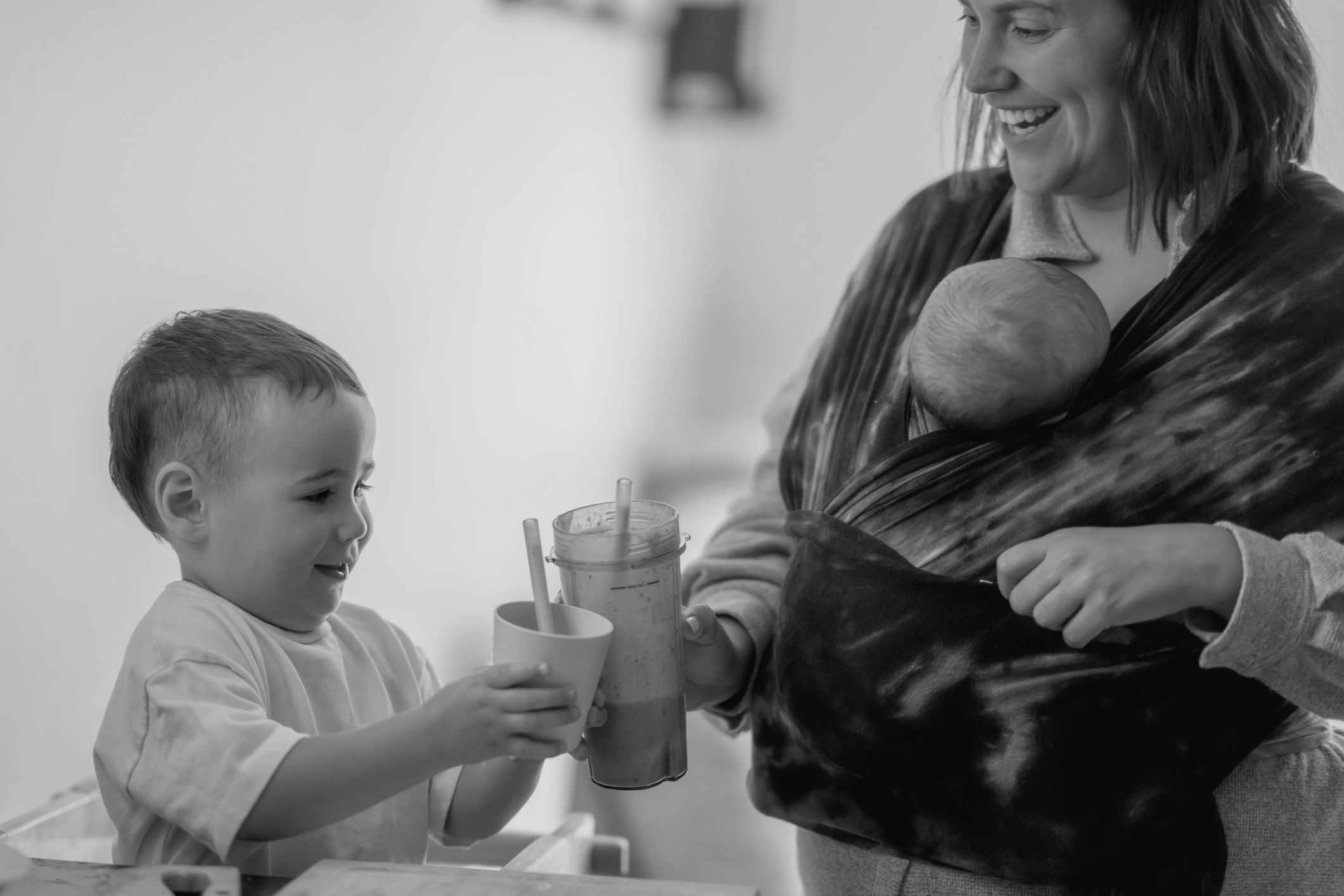 A mother with a baby shares a smoothie with her smiling toddler.
