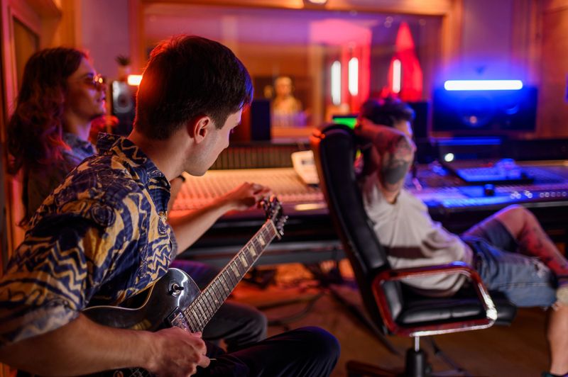 Diverse band member playing electric guitars during rehearsal or recording in a modern music studio with neon illumination. Producer is sitting at the soundboard and singer is in a soundproof booth.