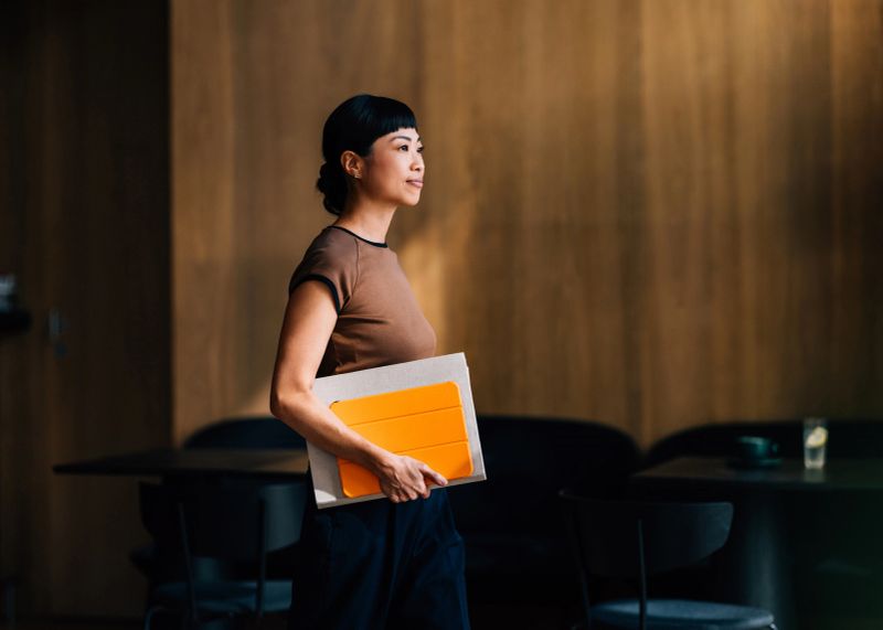 A confident woman walks in a modern office while holding a bright orange notebook, embodying professionalism and focus in a creative workspace.