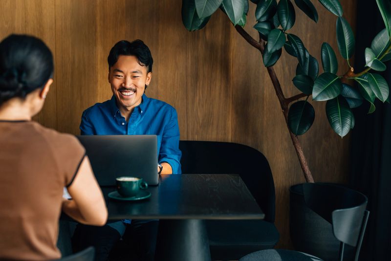 A cheerful man in a casual blue shirt works on his laptop at a cozy coffee shop, engaging in a friendly conversation with a colleague. The atmosphere is warm and inviting.