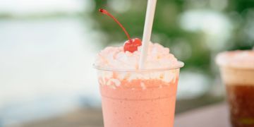 A pink milkshake with whipped cream and a cherry on top in a clear plastic cup.