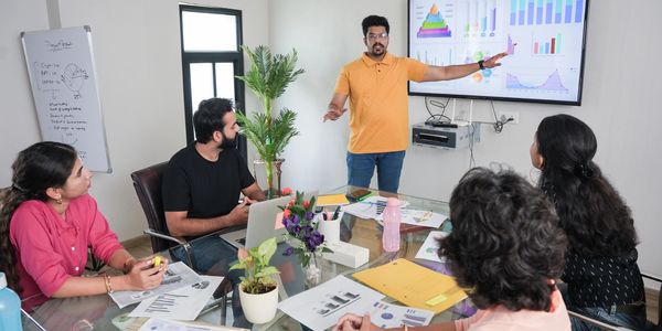 A man in a yellow shirt presents data charts to colleagues in a meeting room.