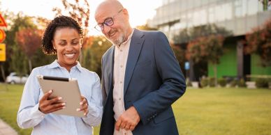 Two colleagues smiling while looking at a tablet outdoors.