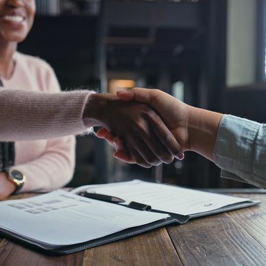 Two people shaking hands over a table with documents and a pen.