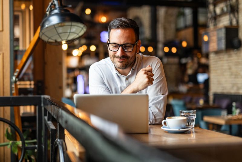 A businessman is sitting at a table in a modern cafe, enjoying a cup of coffee next to him, while his laptop is open and having an online conversation.