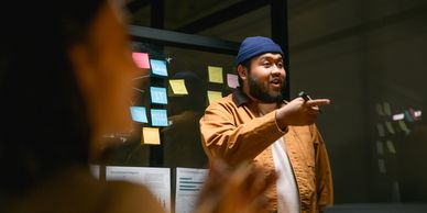 Man in a beanie discussing ideas in front of a glass board with sticky notes.