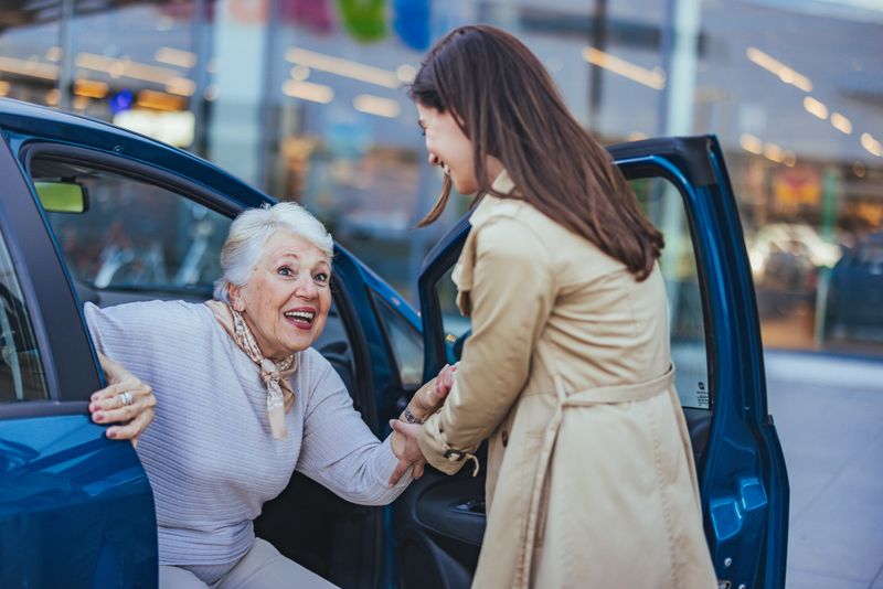 Elderly woman joyfully being helped out of a car by a young woman. The scene depicts care, support, and happiness in an outdoor setting.