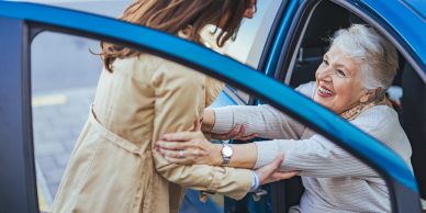 Woman helping elderly lady out of car with a smile.