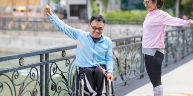 A man in a wheelchair and a woman exercising with resistance bands outdoors.